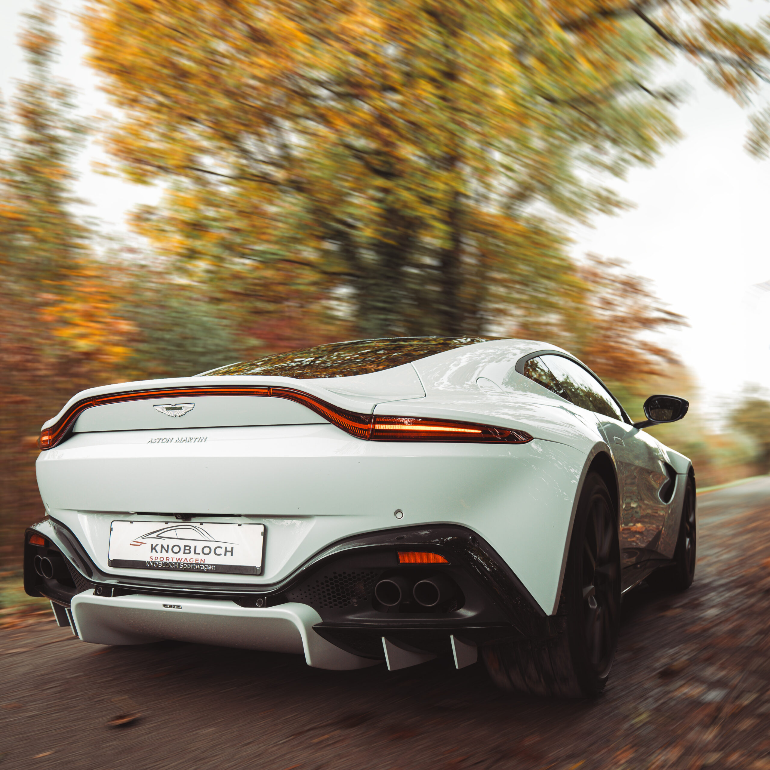 weißer Aston Martin fährt auf einem Waldweg im Herbst
